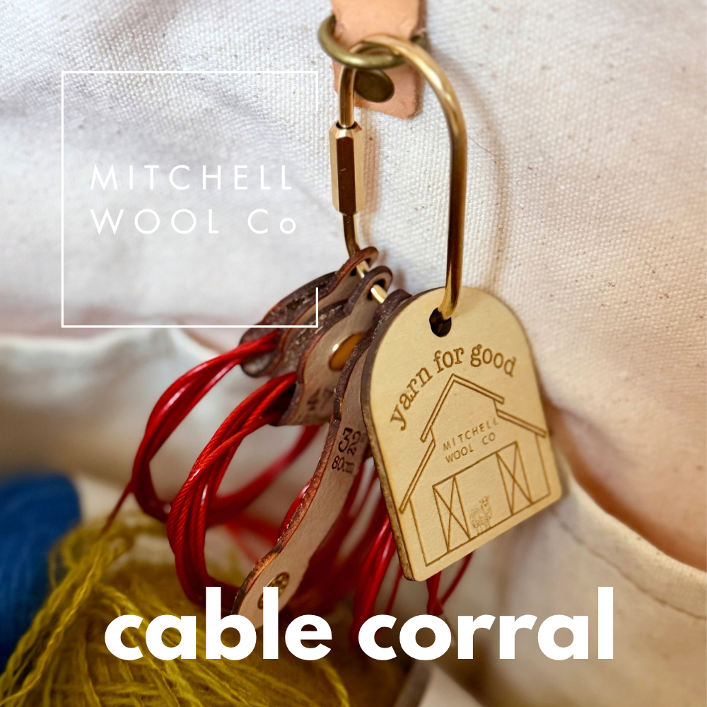 Close-up of a leather cable corral organizer with metal clips and engraved wooden tags, shown hanging against a white fabric background. The product includes red and yellow cable ties, and the text 'MITCHELL WOOL Co' and 'cable corral' is visible on the image.
