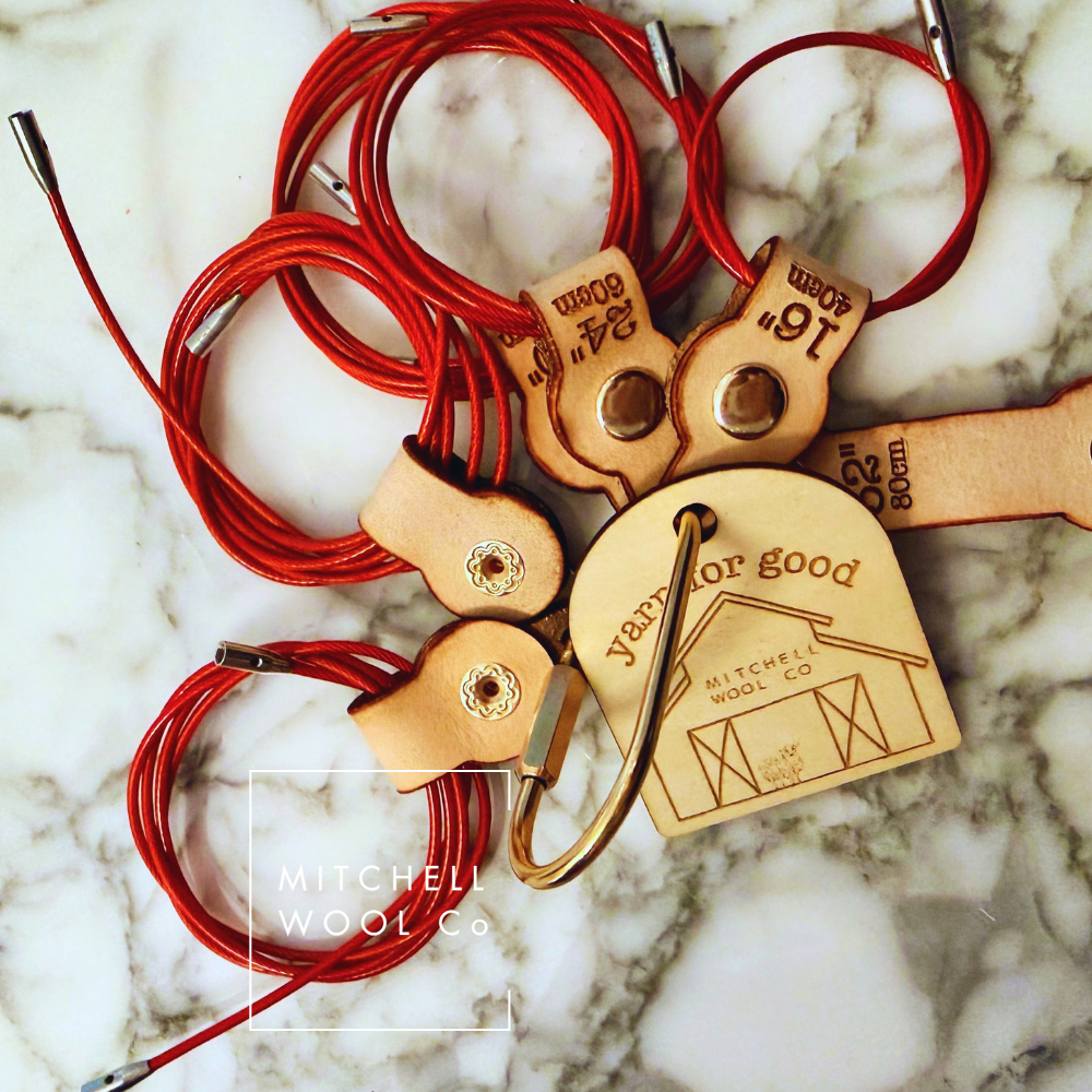 Red wool cable lock with tan leather keeper and branded wooden tag on a white marble background. The lock has engraved size markings and the tag reads 'Yor for good MITCHELL WOOL CO.
