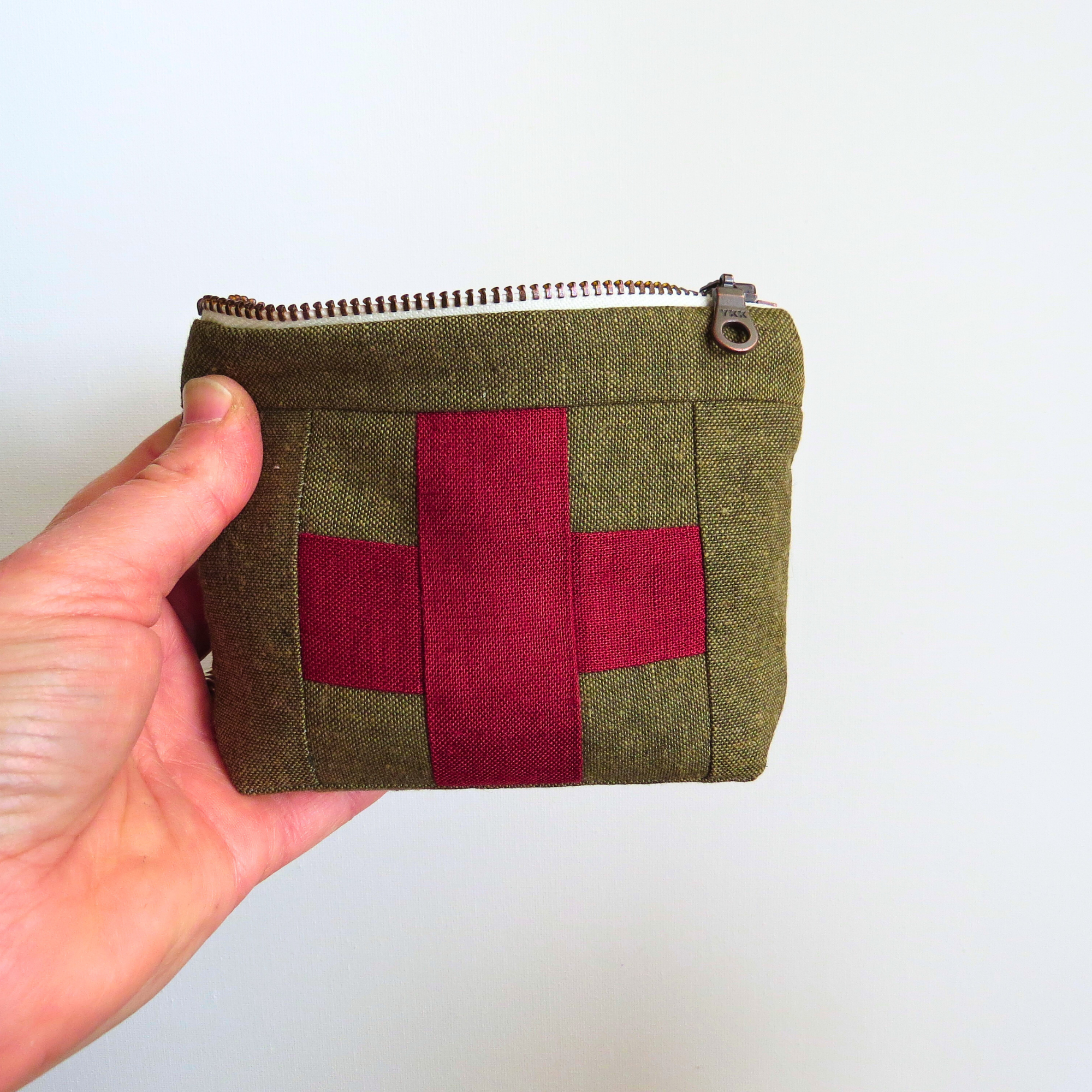 Hand holding a small green fabric pouch with a red cross design on a white background