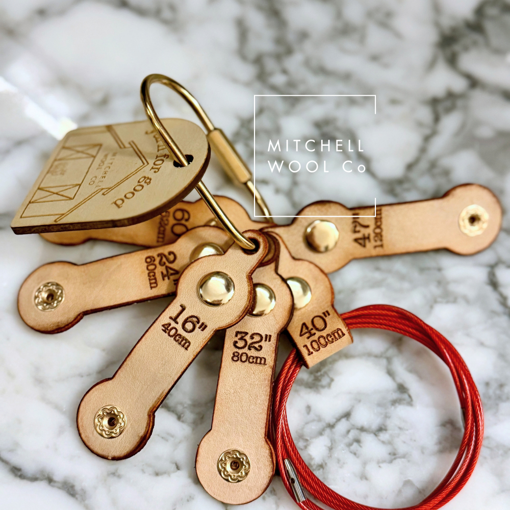 A set of wooden knitting needle size cards with brass metal rings and a red yarn sample, displayed on a white and gray marble surface. The brand name 'Mitchell Wool Co.