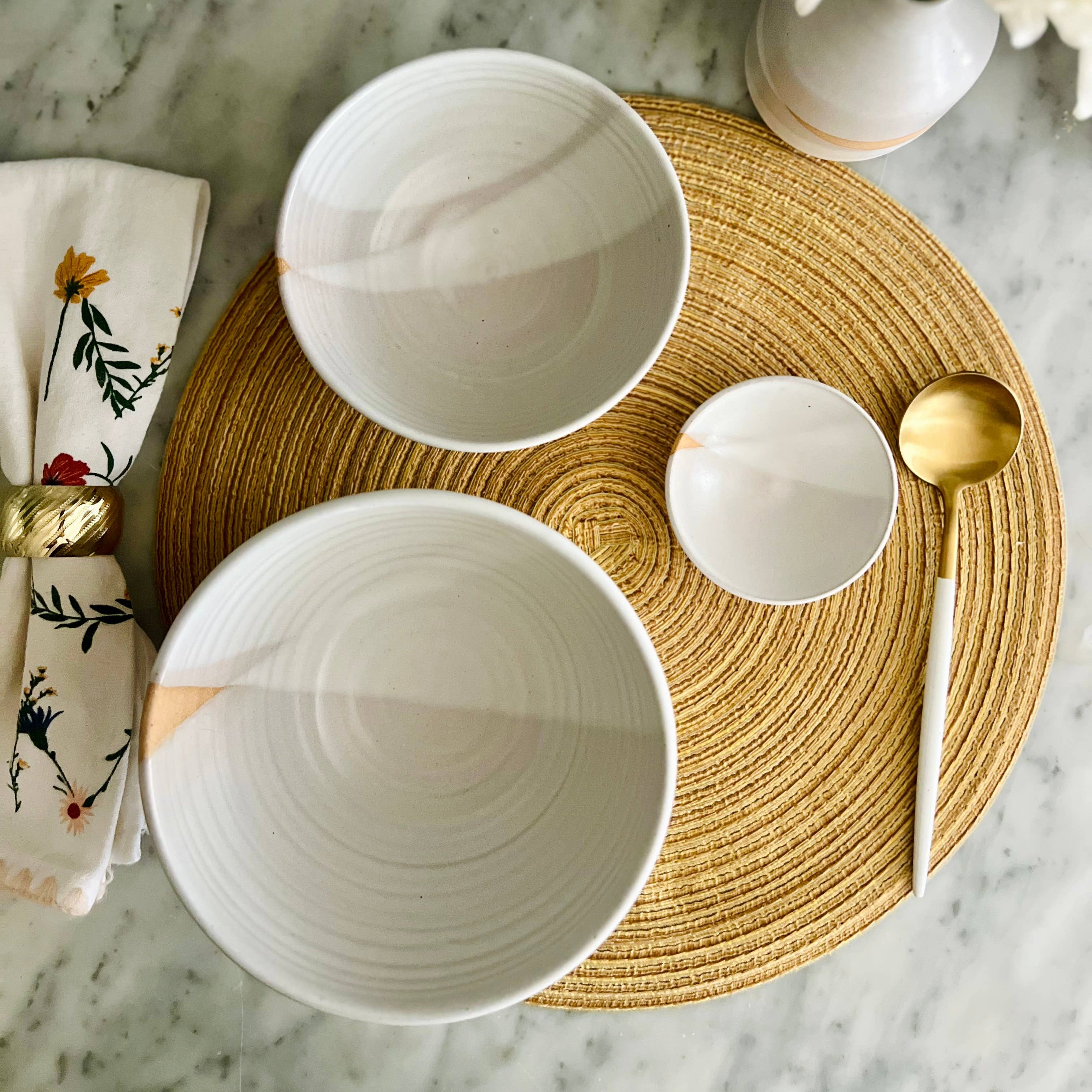 Two white ceramic bowls on a woven placemat with a gold spoon and floral napkin on a marble surface.