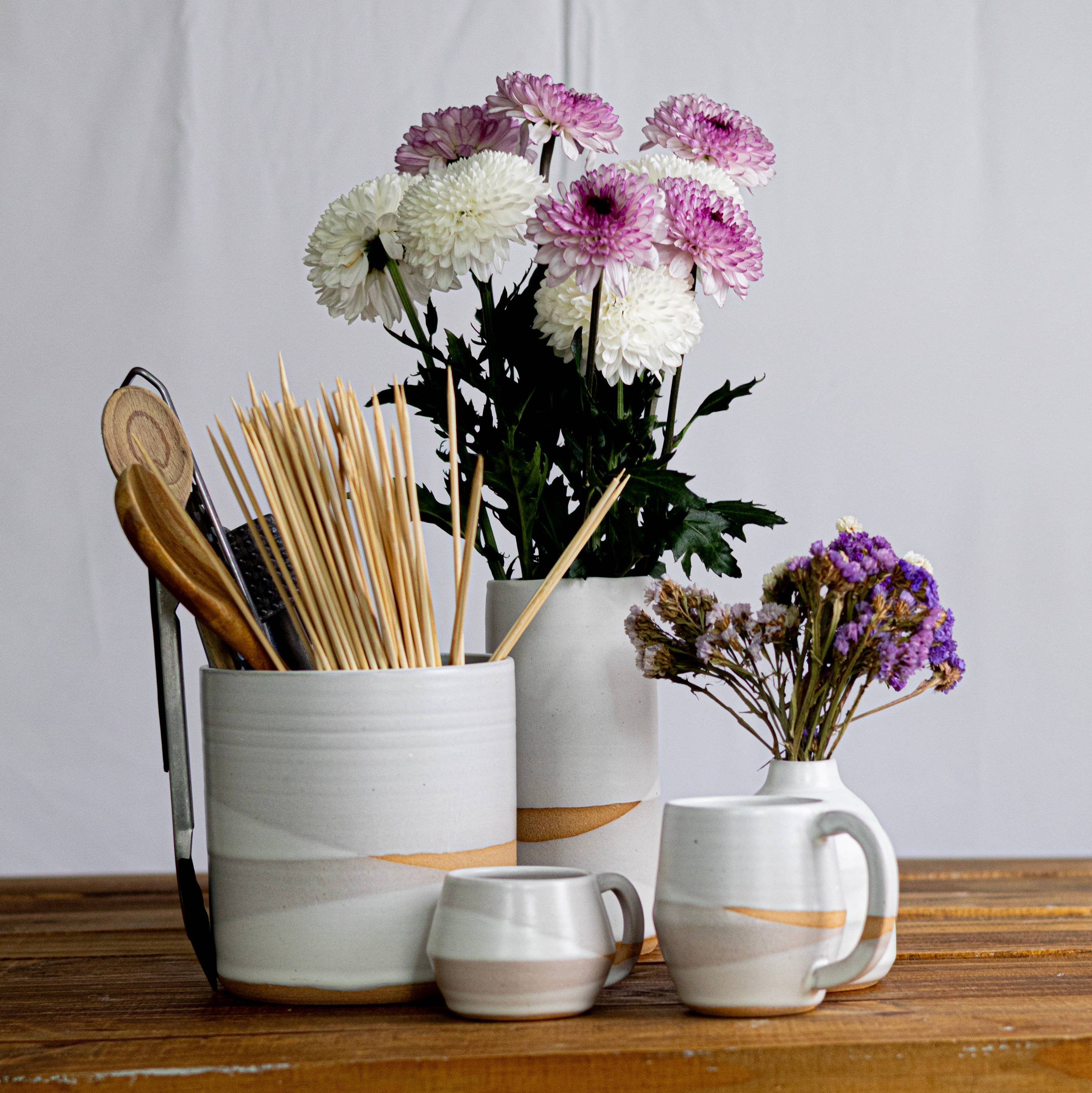 White ceramic containers with gold accents on a wooden surface, containing flowers and kitchen utensils.