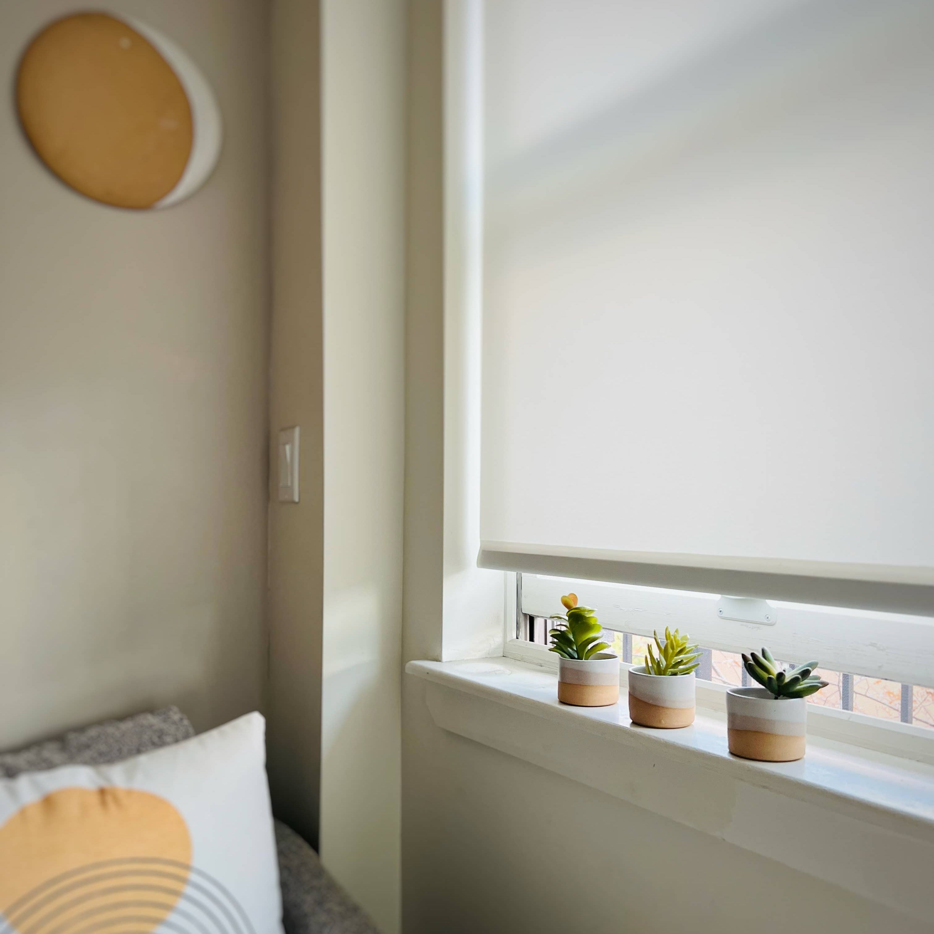 Small indoor garden with three potted plants on a windowsill next to a bed with decorative pillows.