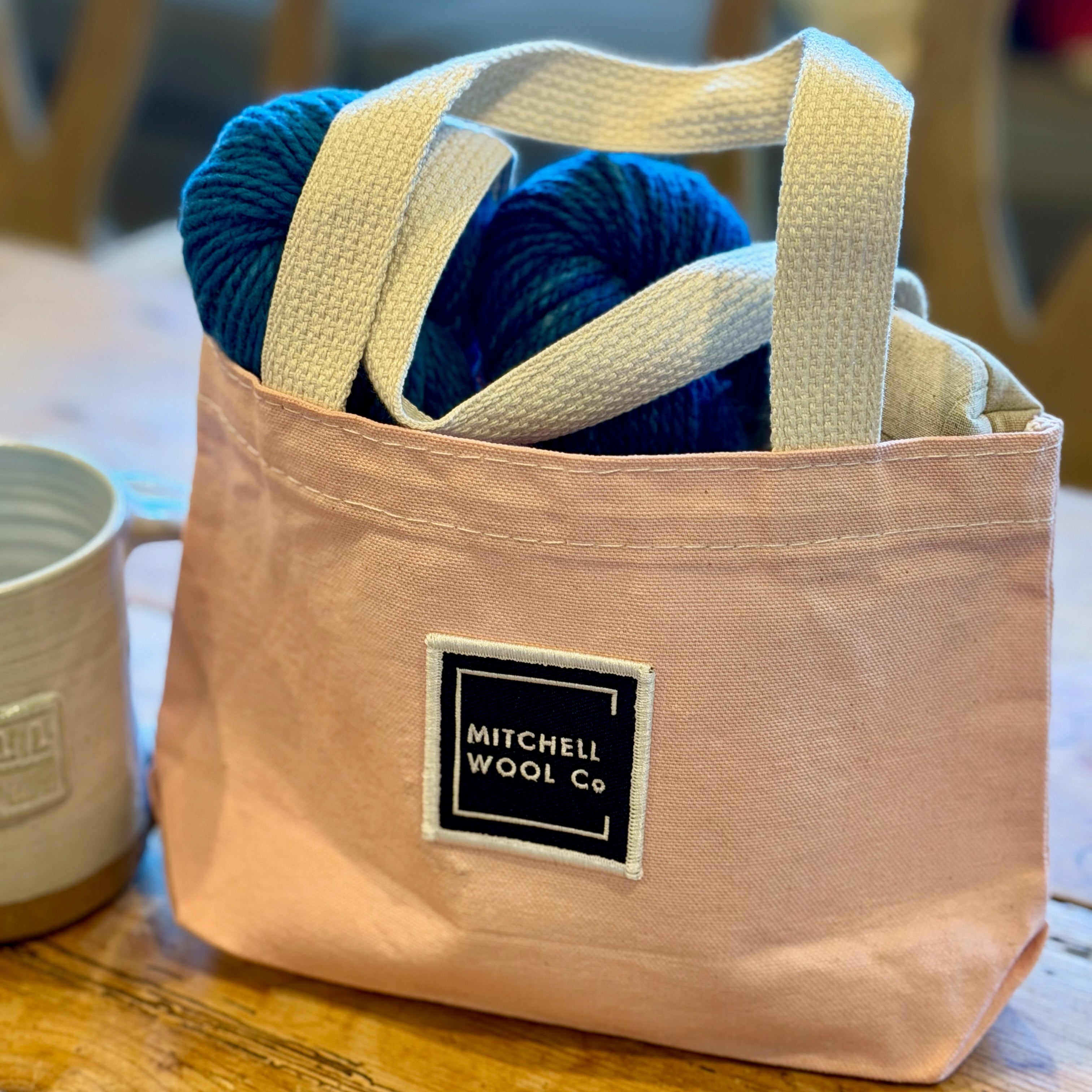 Pink tote bag with yarn and a mug on a wooden table, featuring Mitchell Wool Co. branding.