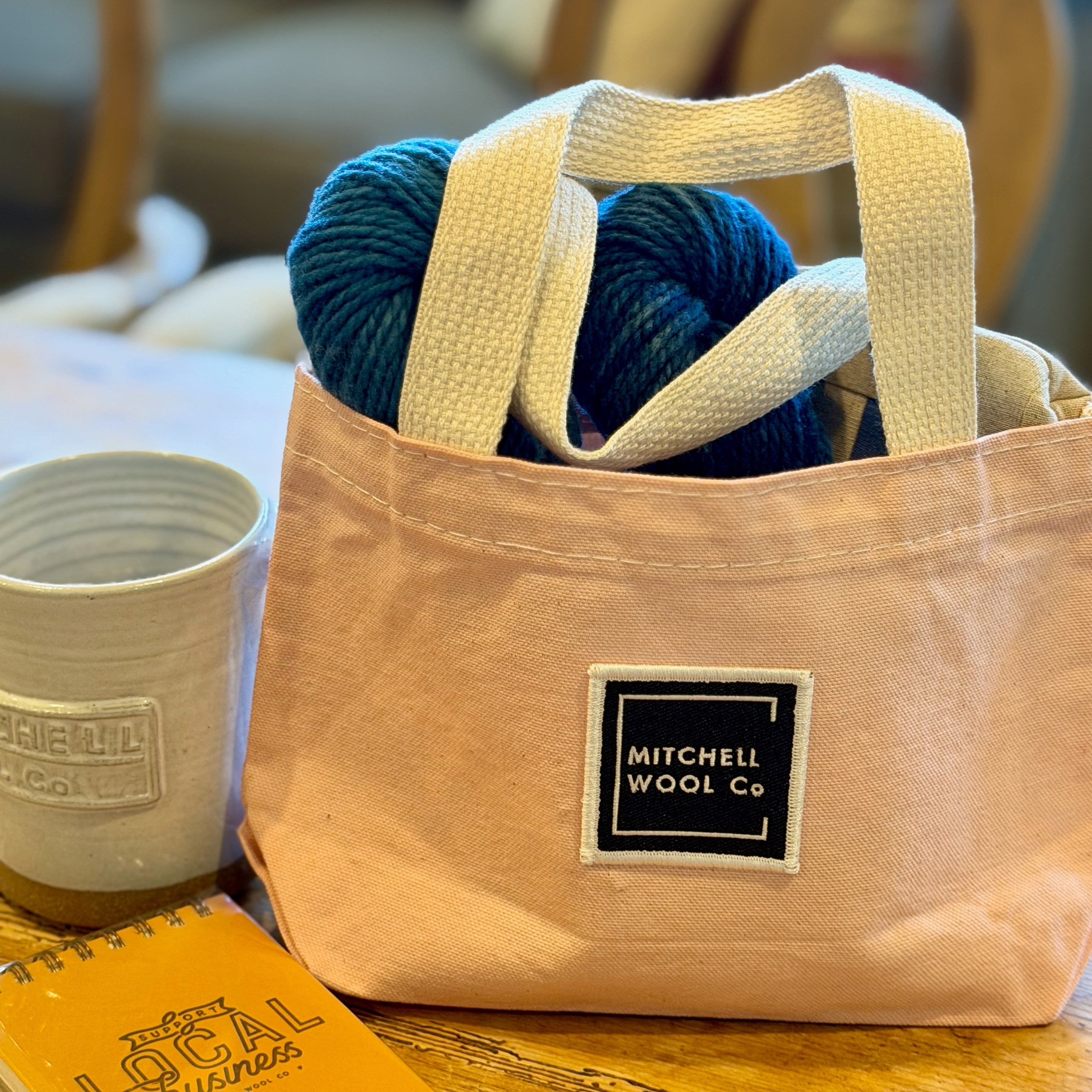 Blush pink tote bag with blue yarn, a mug, and notebook on a wooden table.