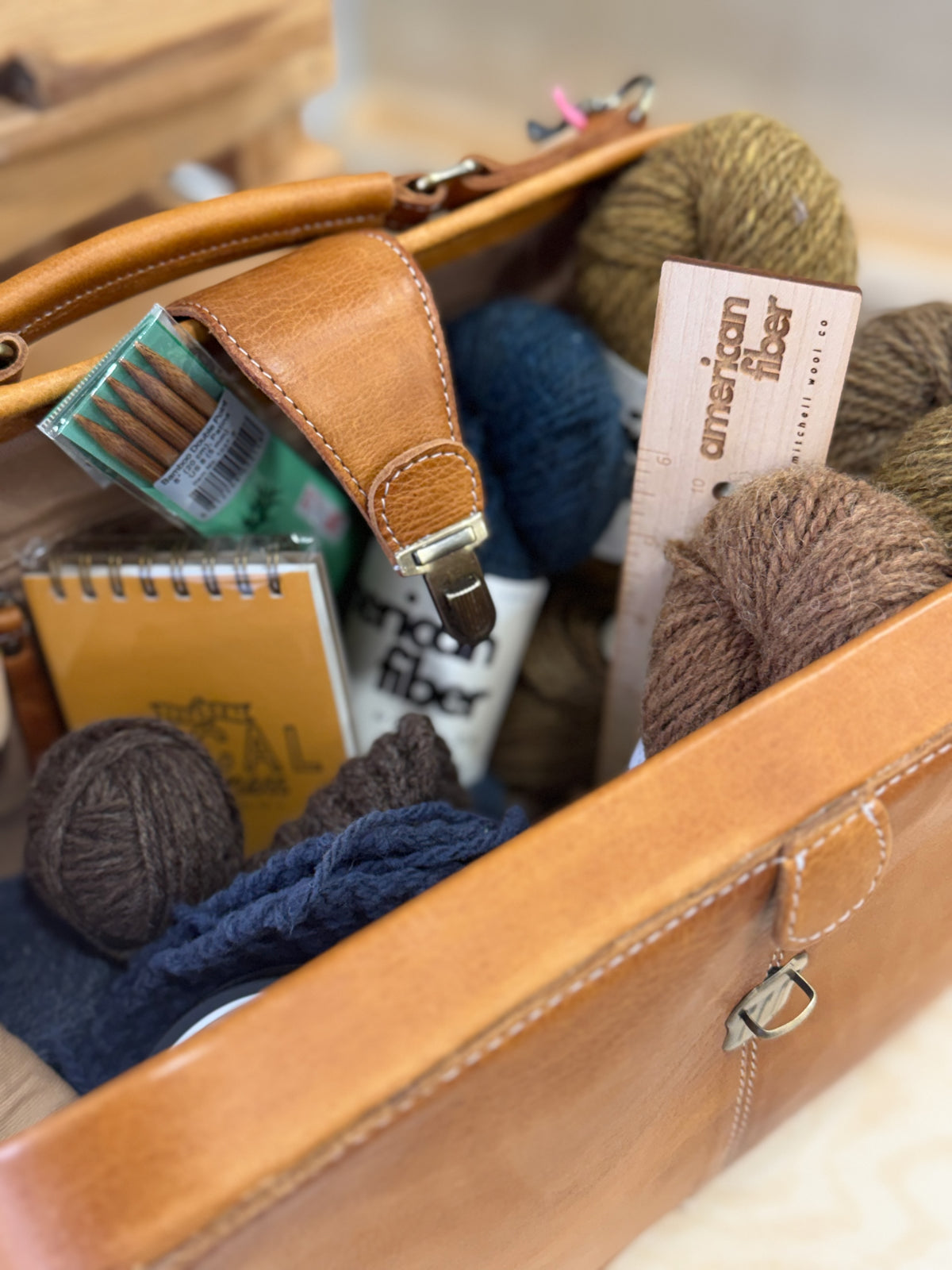 A tan leather organizer tote filled with knitting supplies, including balls of yarn in brown, navy, and olive, a notebook, a small box of knitting needles, and a card reading 'american fiber'.