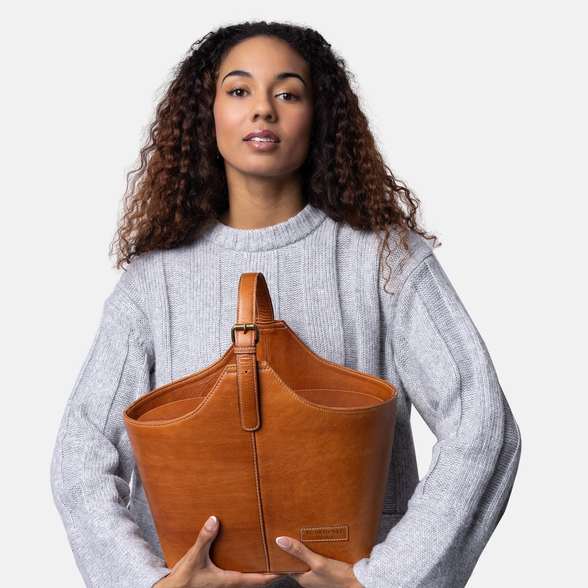 Woman with light brown skin  & a grey sweater holding a brown leather bag against a white background