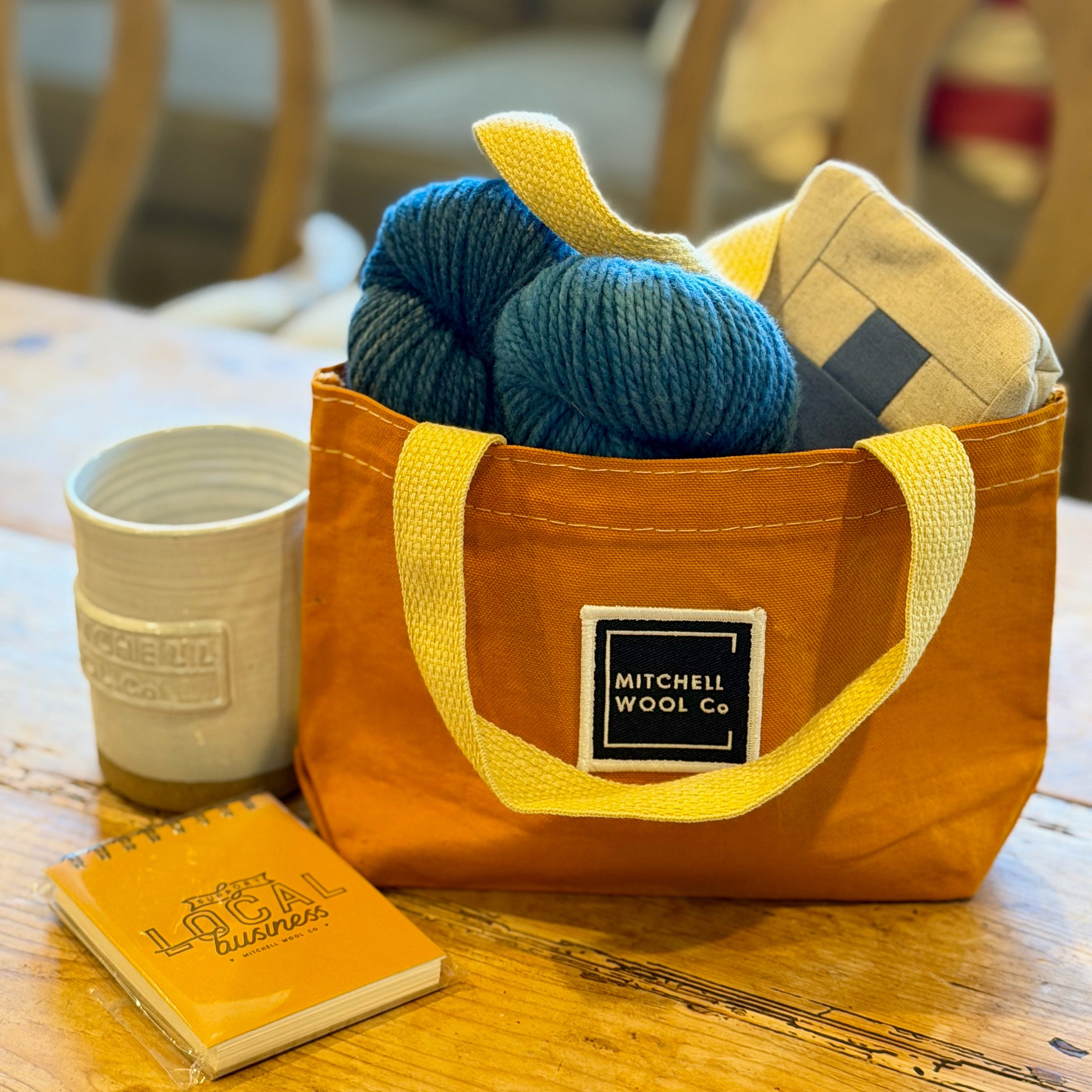 Orange bag with yarn and knitting needles on a wooden table, with a notebook and cup in the foreground.