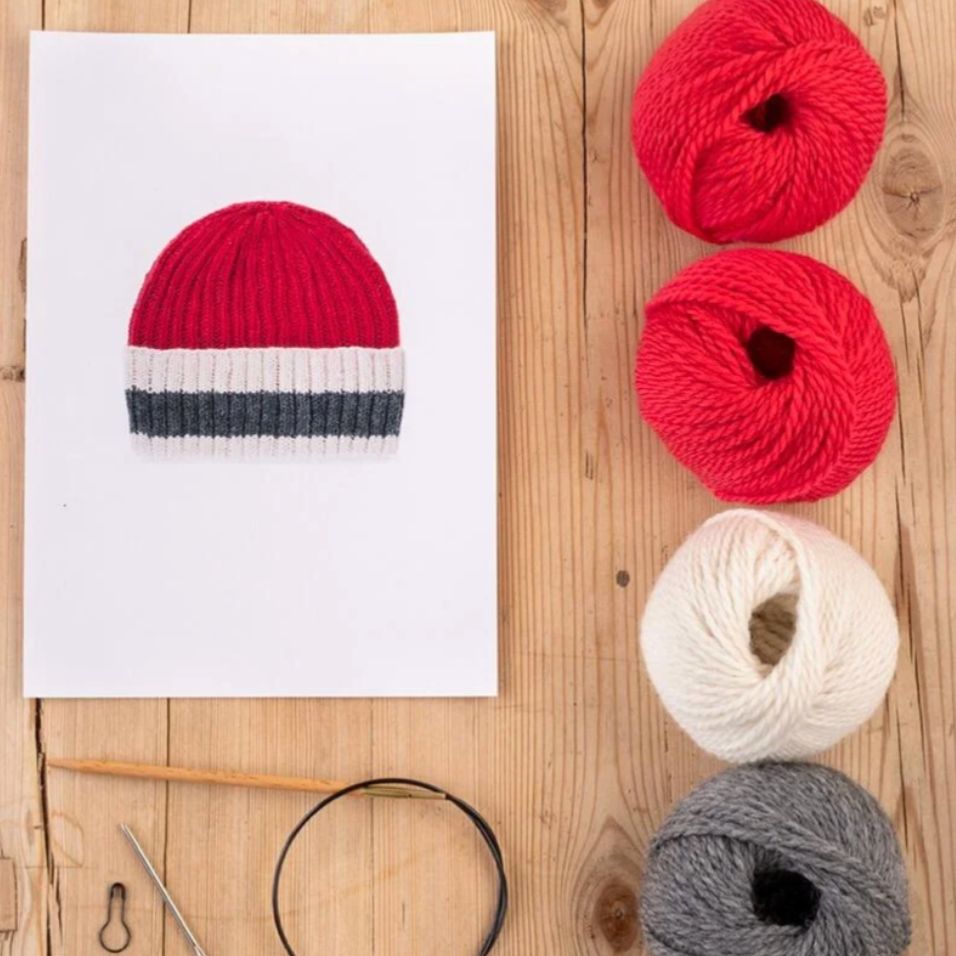 Red and white striped knitted hat with yarn balls and knitting tools on a wooden surface