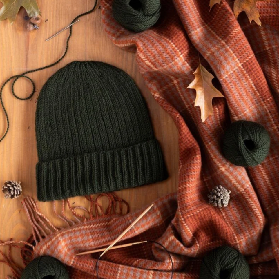 Black knit beanie with green yarn on a wooden surface with a plaid blanket and autumn leaves.