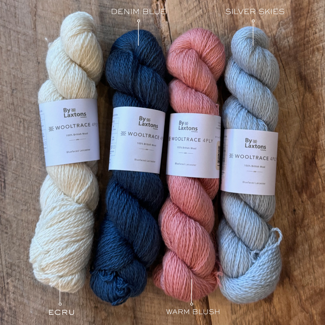 Four skeins of yarn in ecru, denim, warm blush & silver skies with labels on a wooden surface