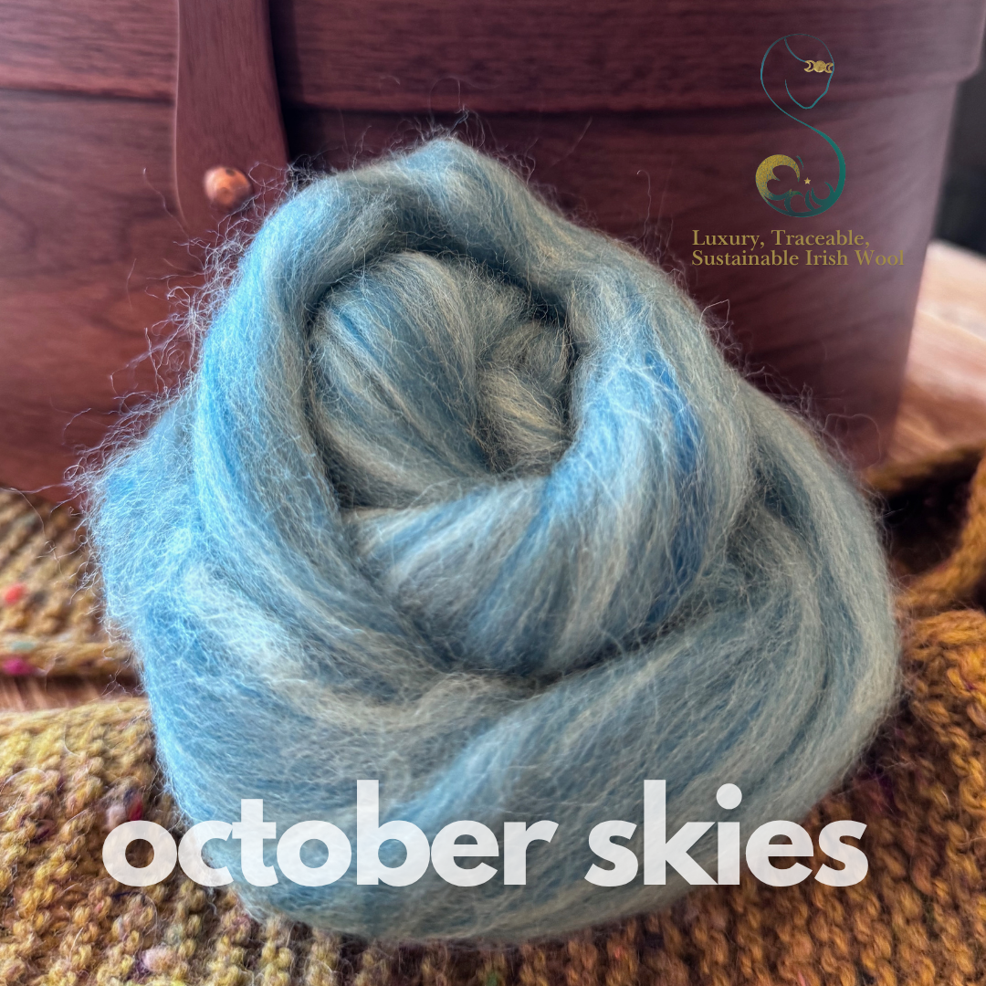 Light turquoise & grey marled dollup of combed top from BFL Irish sheep labeled 'October Skies' on a textured surface with a wooden box in the background.