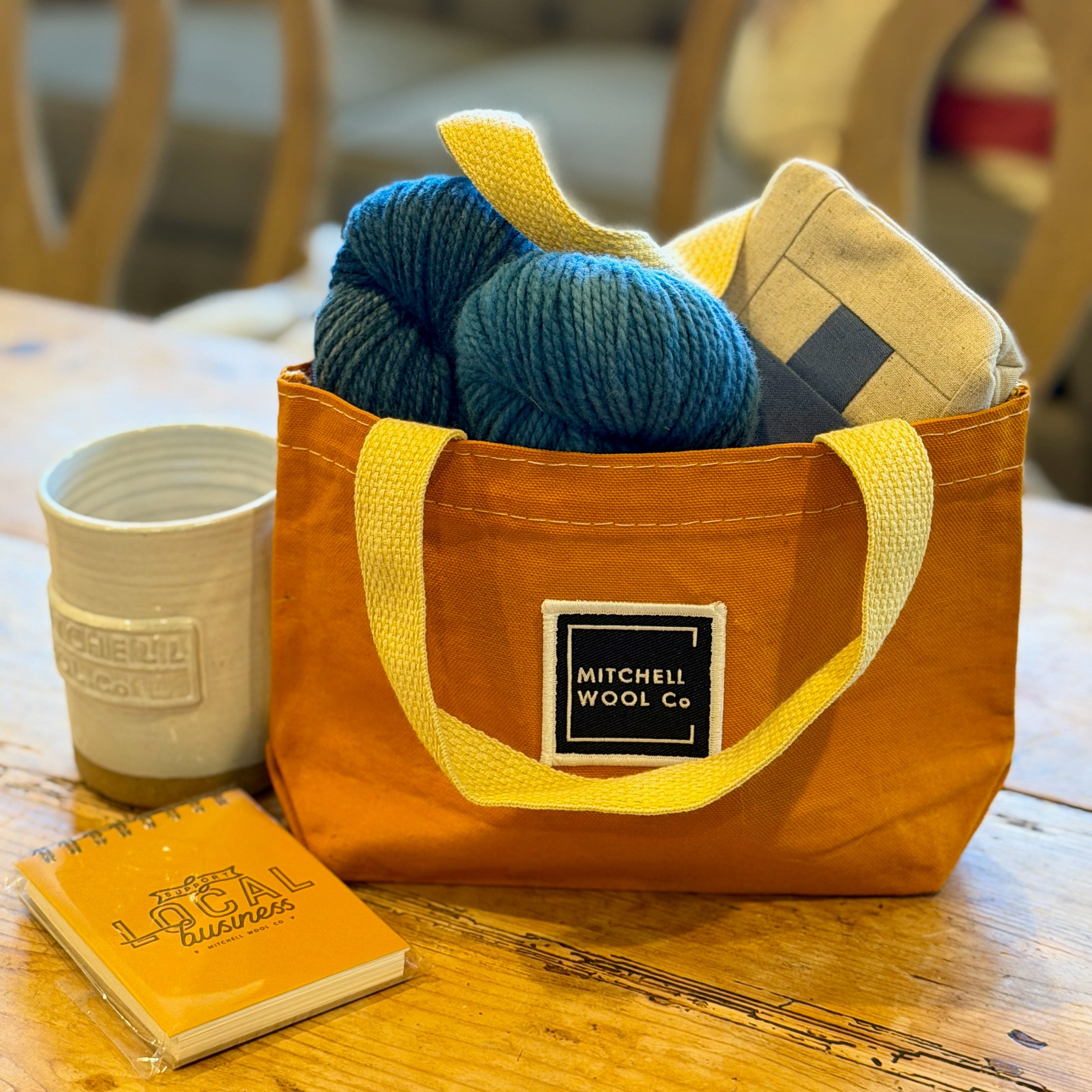 Orange bag with yellow straps containing yarn and a notebook on a wooden table.